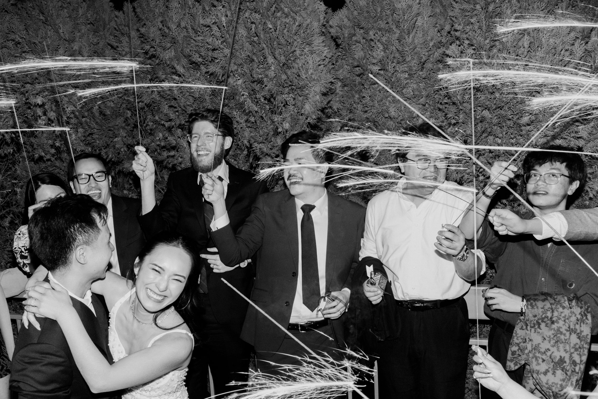 Black and white photo of bride, groom and guests holding sparklers during joyful wedding send-off.
