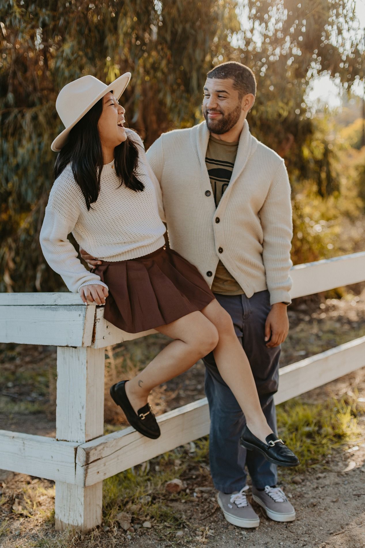 A couple in cozy sweaters sits together on a white wooden fence during a golden autumn afternoon at Will Rogers State Park.