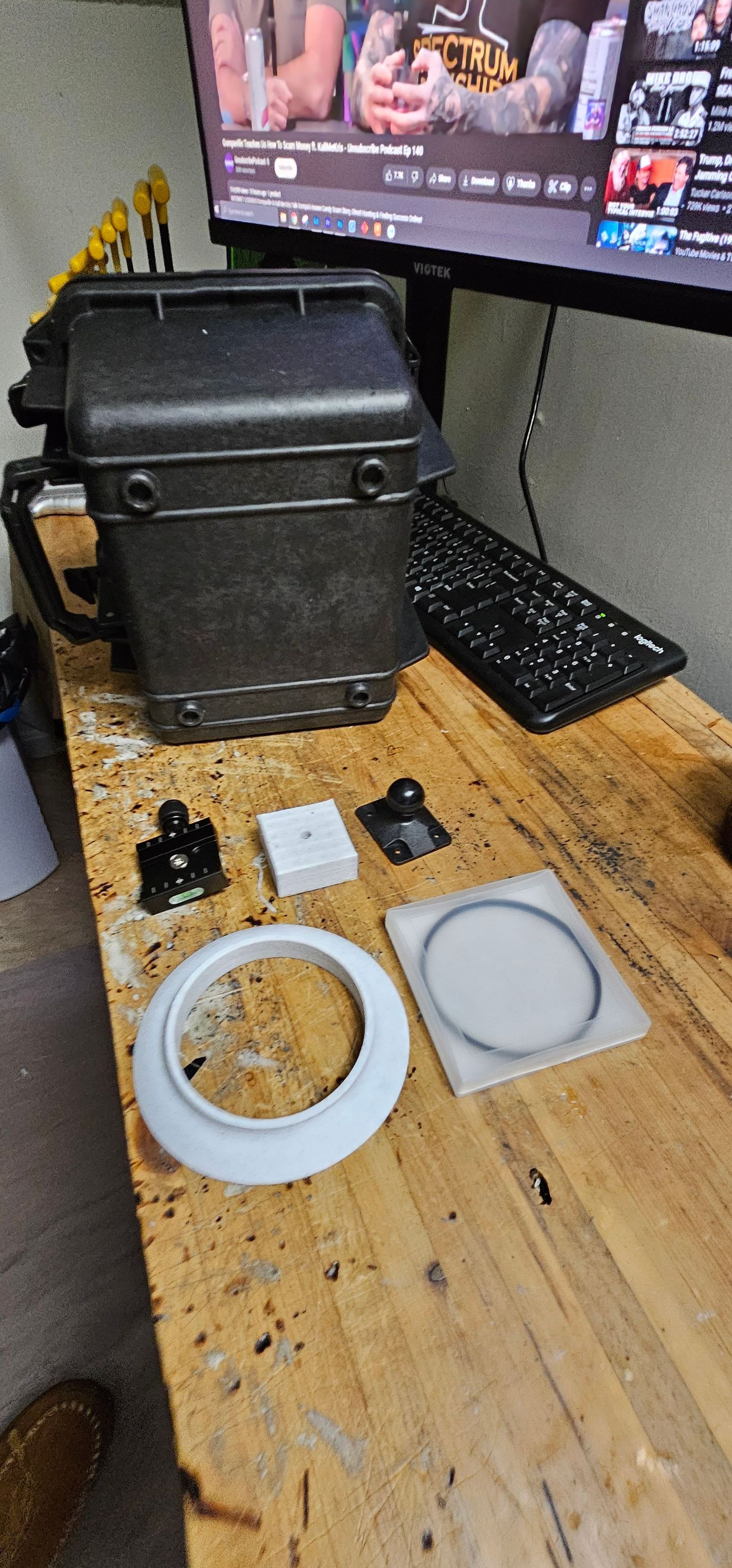 Black hard case with white lens ring, filter, and mounting hardware on a wooden workbench near a monitor.
