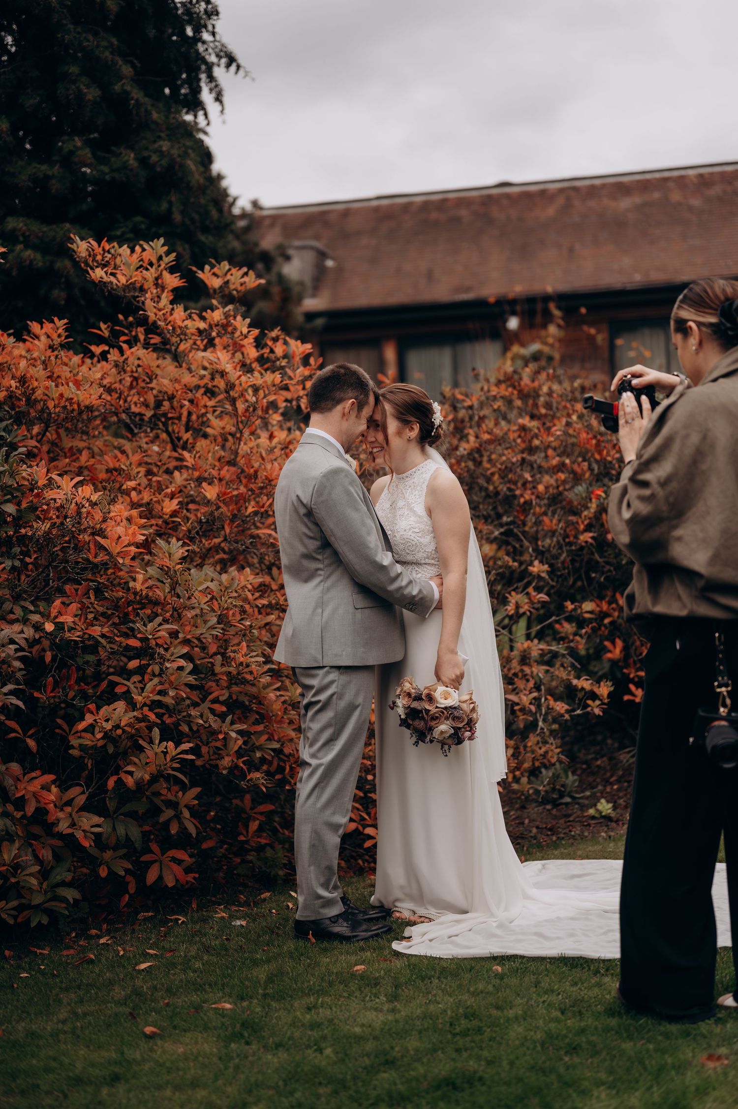 A wedding photographer captures a couple embracing outdoors with vibrant orange autumn foliage in the background.