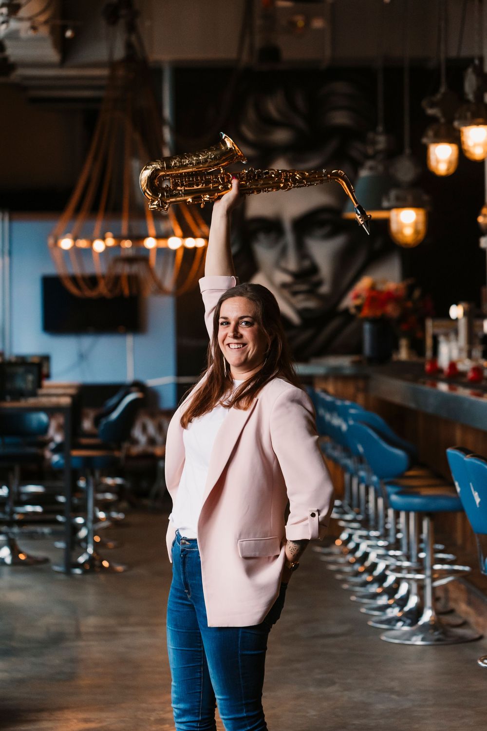 Musician in pink blazer and jeans poses with saxophone in stylish bar with vintage lighting and wall art.