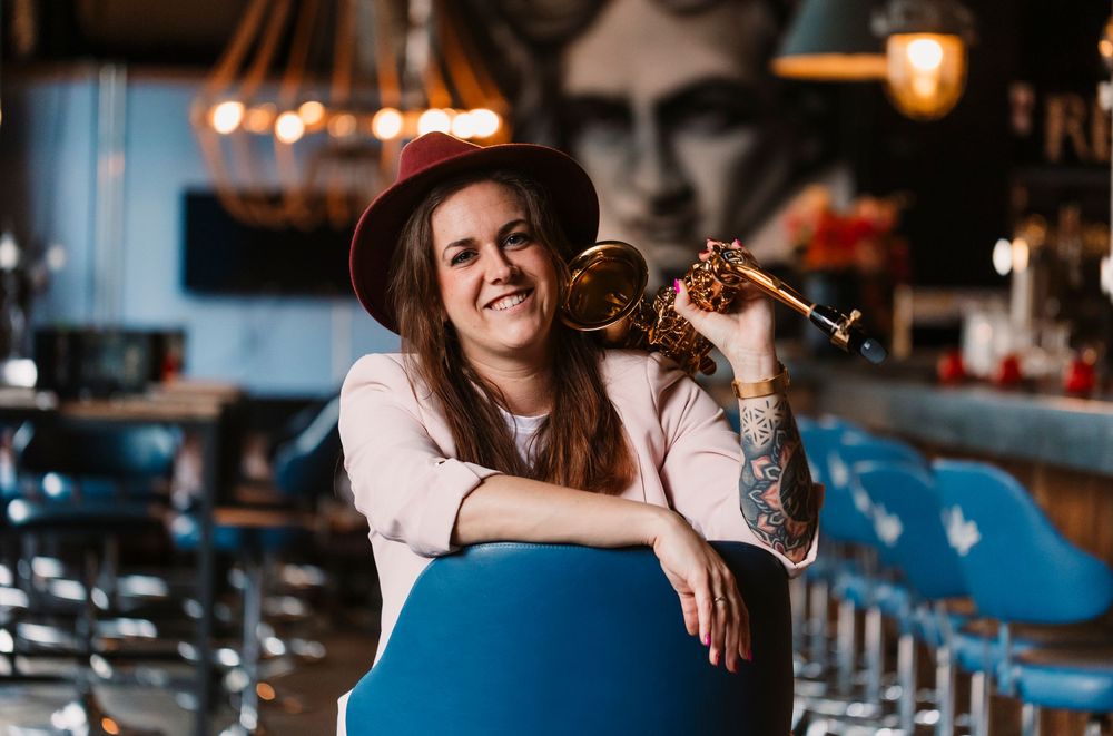 Saxophone player in pink blazer leans on blue bar chair in atmospheric venue.