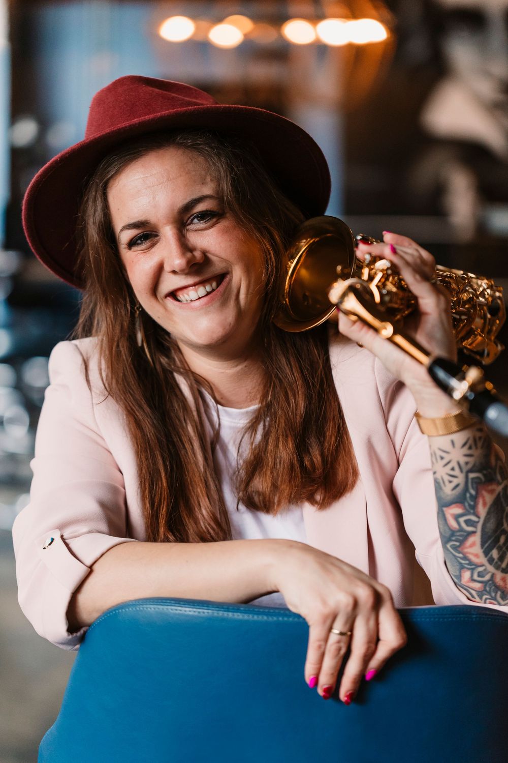 Musician in burgundy hat and pink blazer poses with saxophone in moody bar setting.