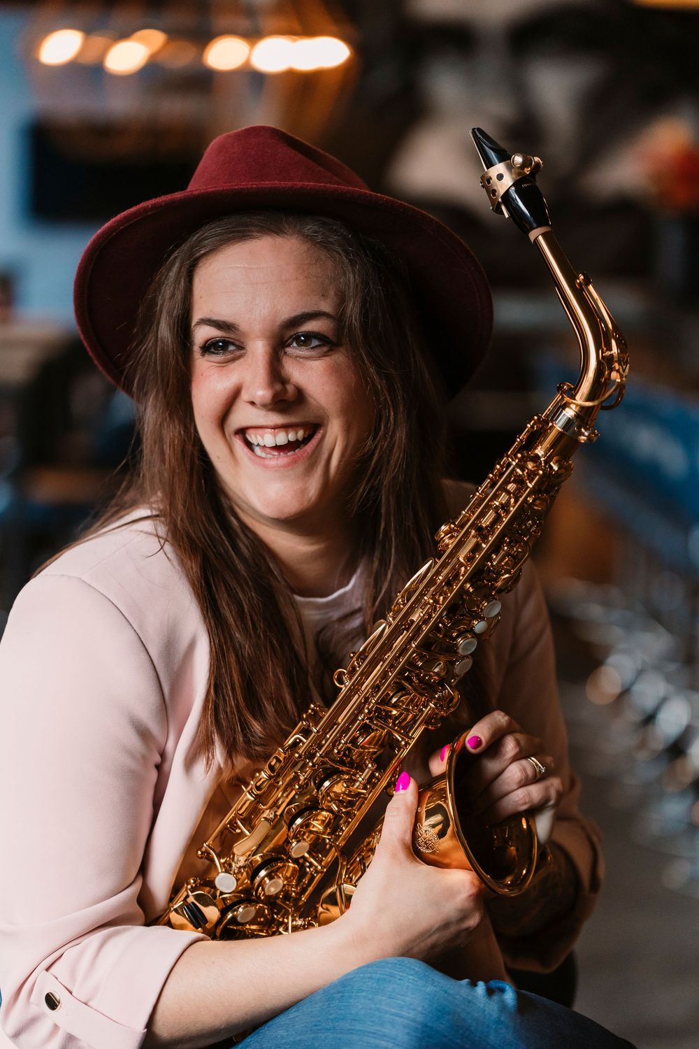 Saxophonist wearing burgundy hat and pink jacket smiles while holding instrument in bar setting.
