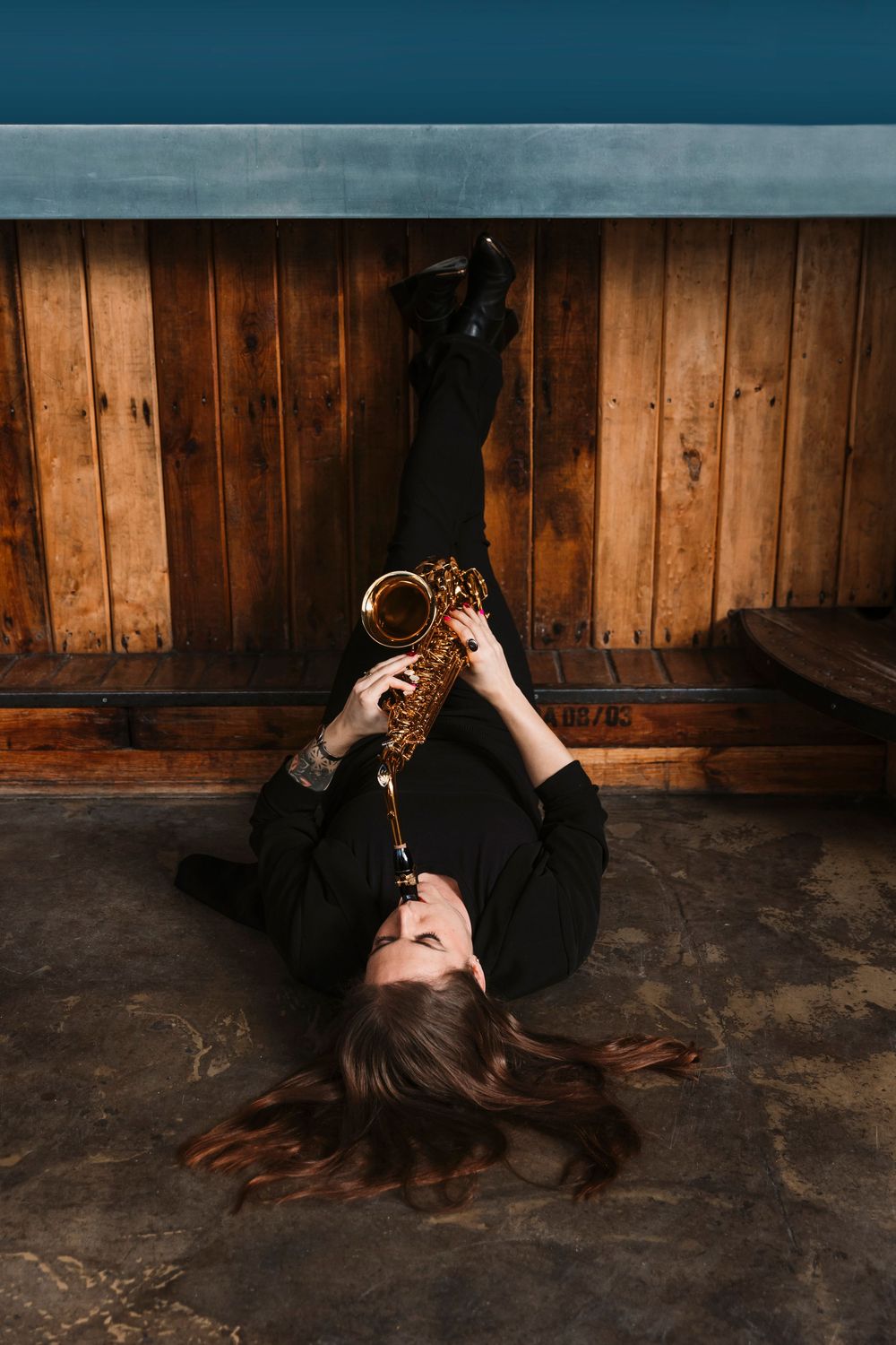 Musician lies on floor holding saxophone against rustic wooden wall backdrop.
