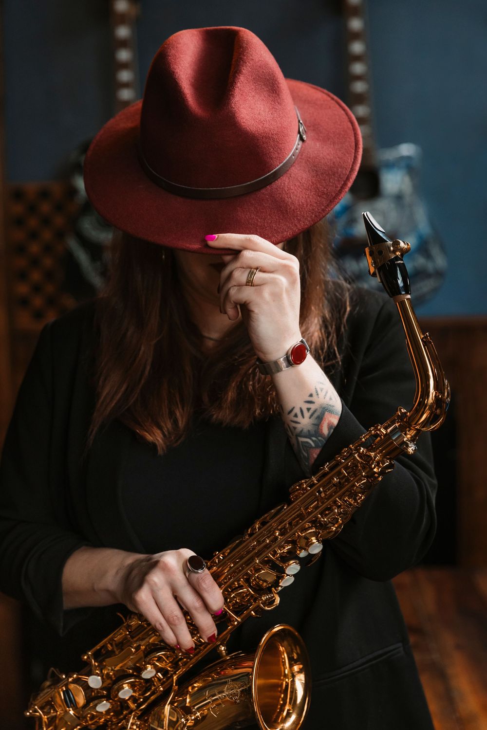 Musician in burgundy hat and black outfit holds saxophone in moody bar setting.