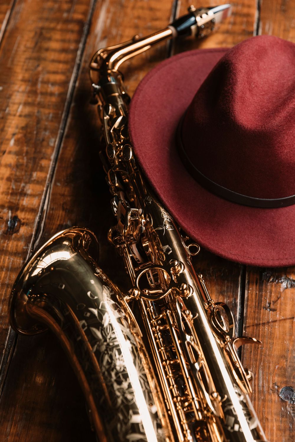 Close-up of a brass saxophone and burgundy fedora hat on wooden surface.
