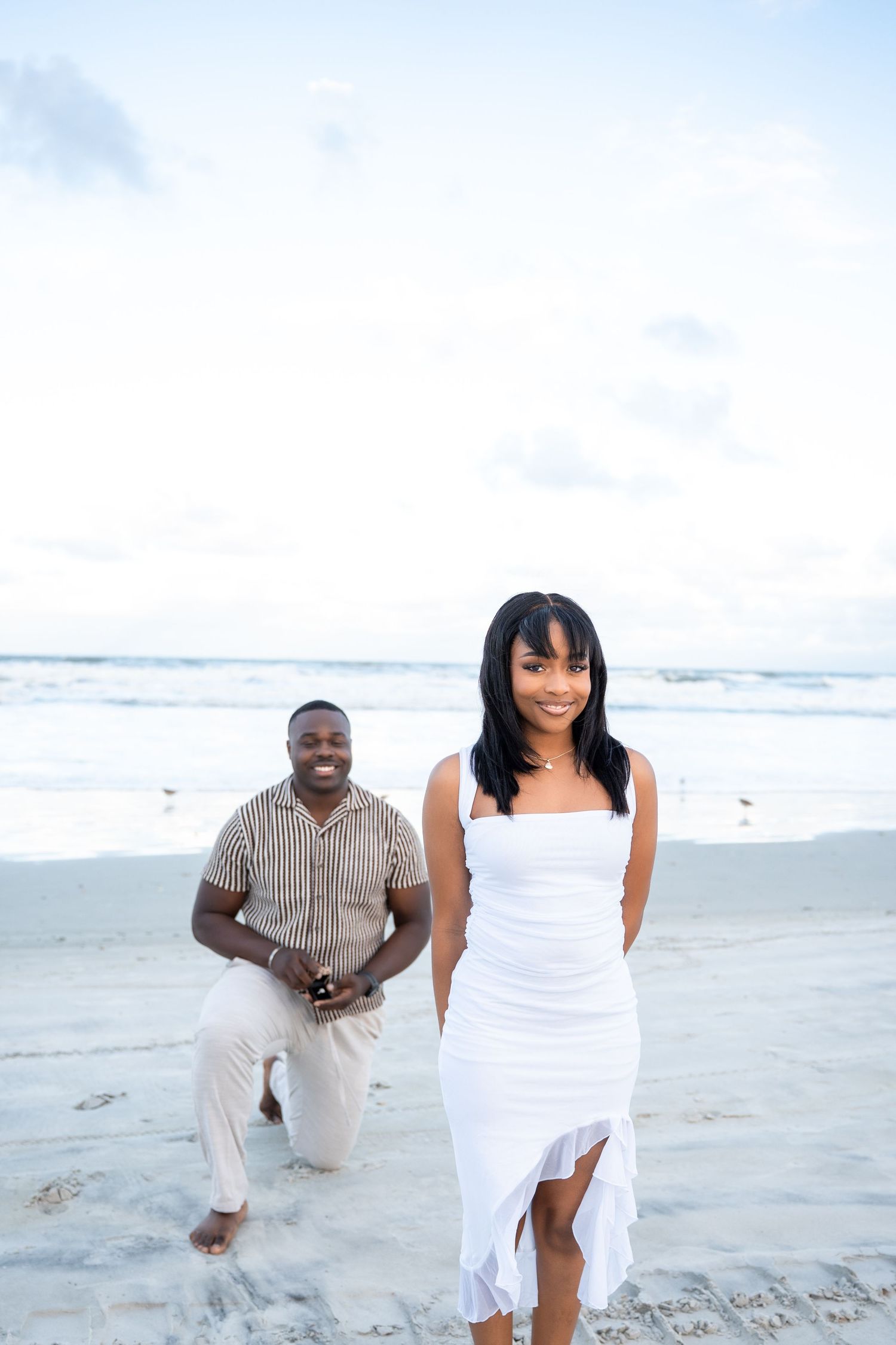 A couple in white outfits playfully runs and poses together on a sunny beach with ocean waves in the background.