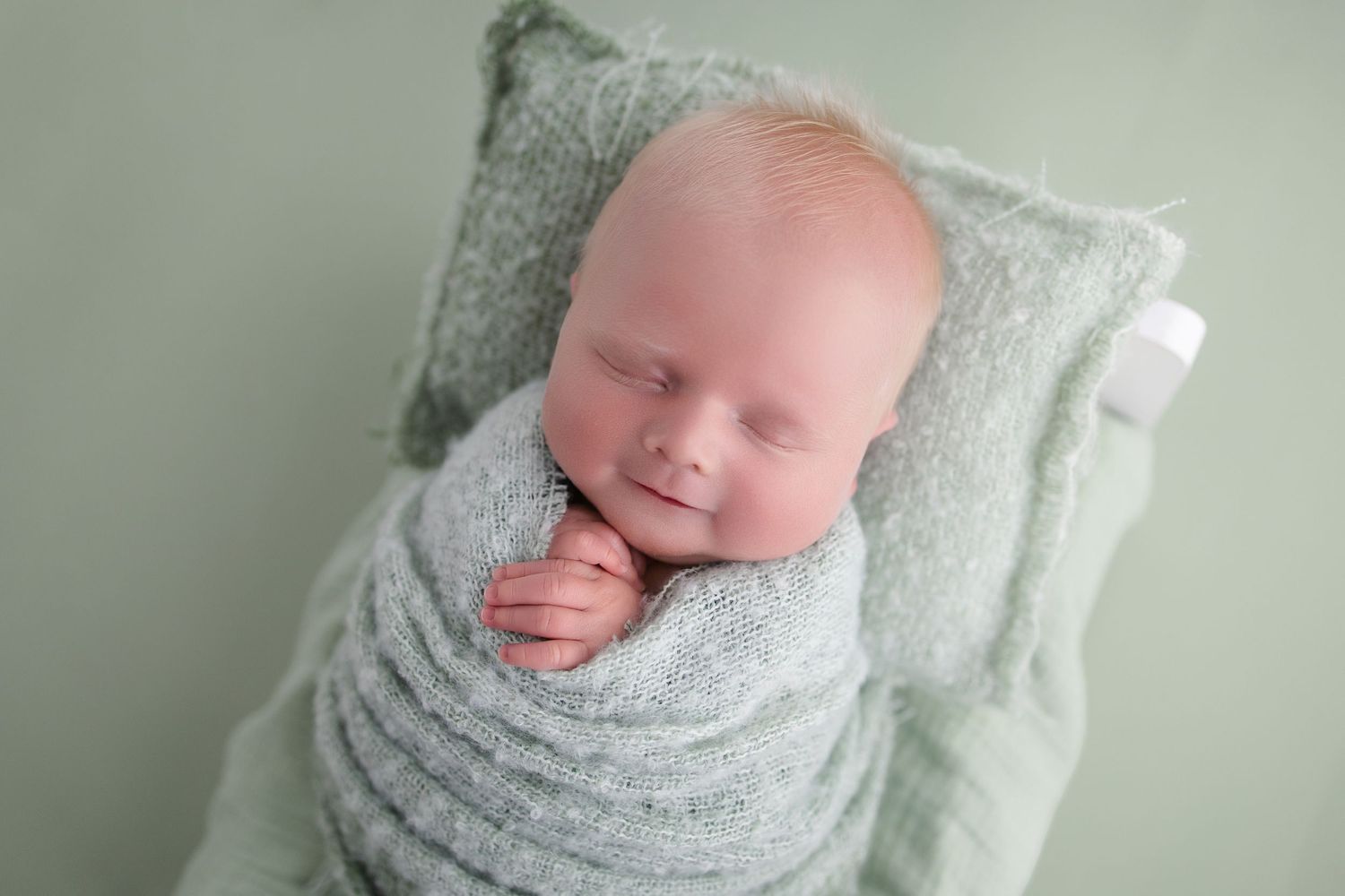 Sleeping newborn baby swaddled in a soft mint green wrap, resting on a tiny matching pillow and bed.
