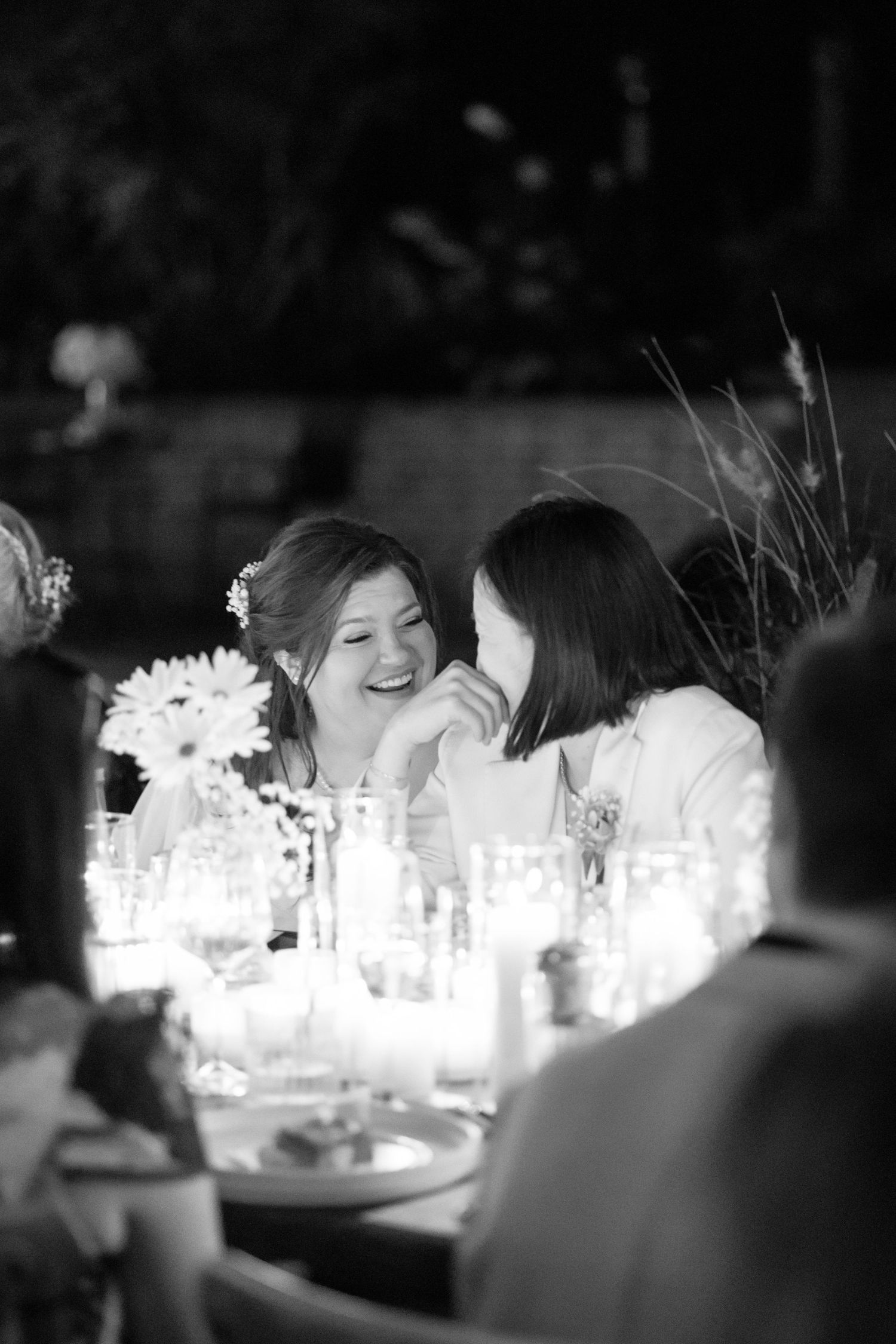 Black and white photo at same-sex wedding of two brides laughing together at a candlelit wedding reception table. Candid Wedding Photography