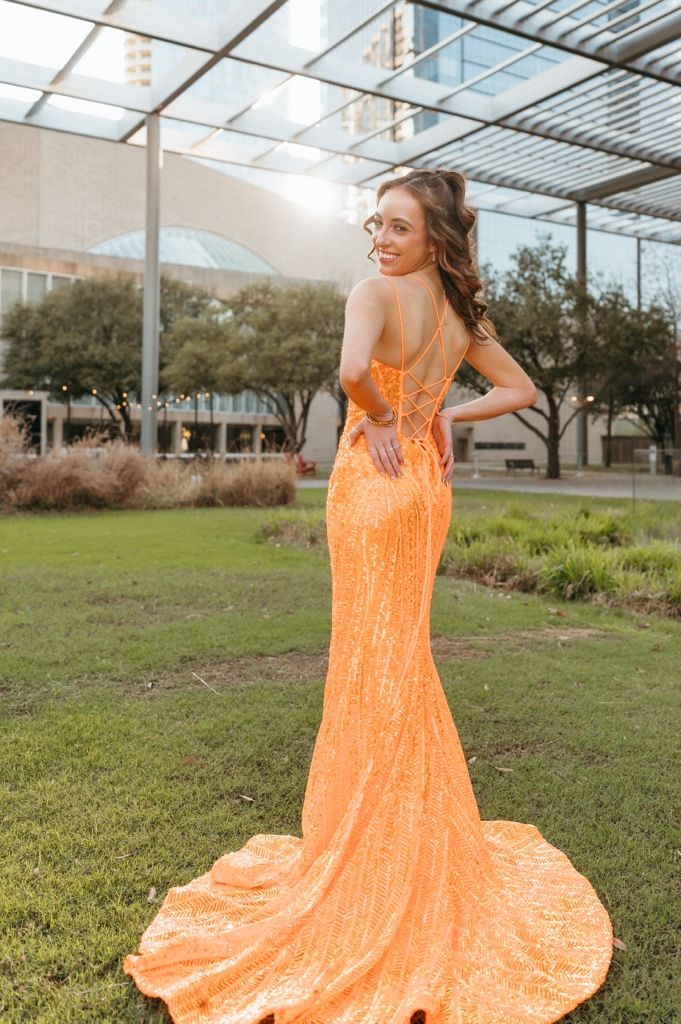 Woman in orange sequin mermaid gown with train poses outdoors near modern building on green lawn.