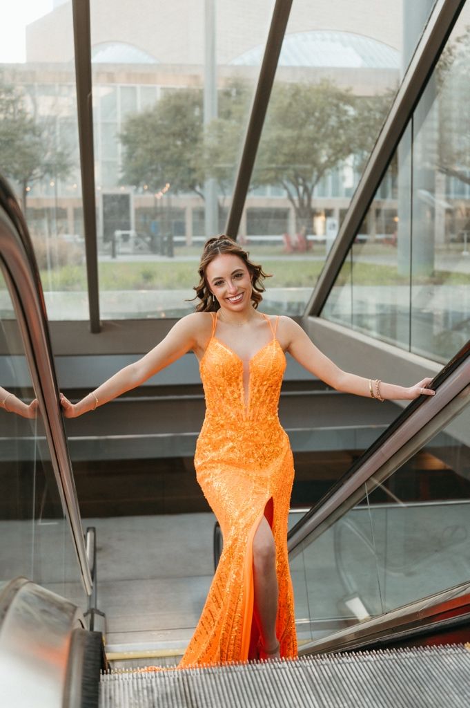Woman in orange sequin gown posing on escalator, smiling confidently in elegant formal dress.