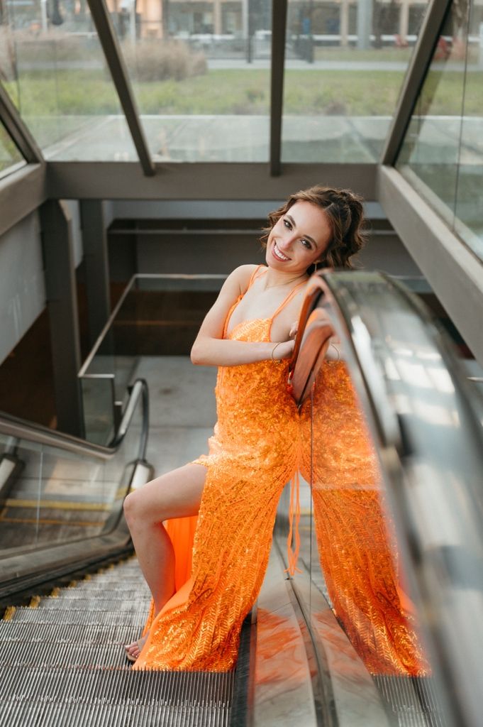 A smiling woman in an elegant orange sequin gown poses gracefully on an escalator in a modern glass building.