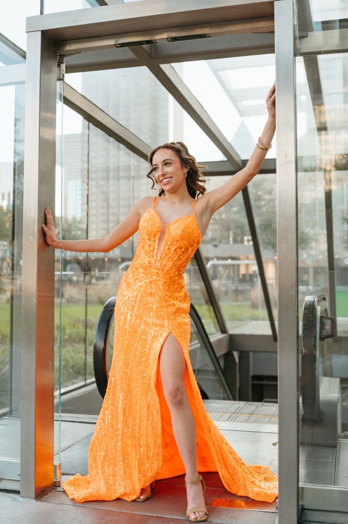 Woman in orange sequin gown with slit poses elegantly in glass elevator doorway, smiling confidently.