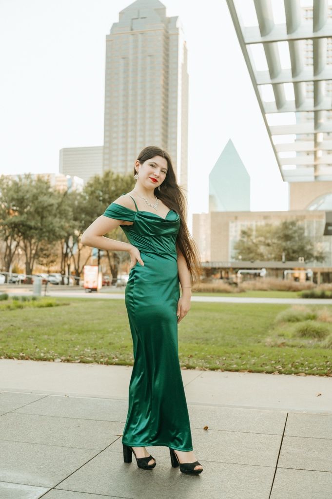 Woman in elegant green satin off-shoulder gown posing outdoors with Dallas city skyline in background.