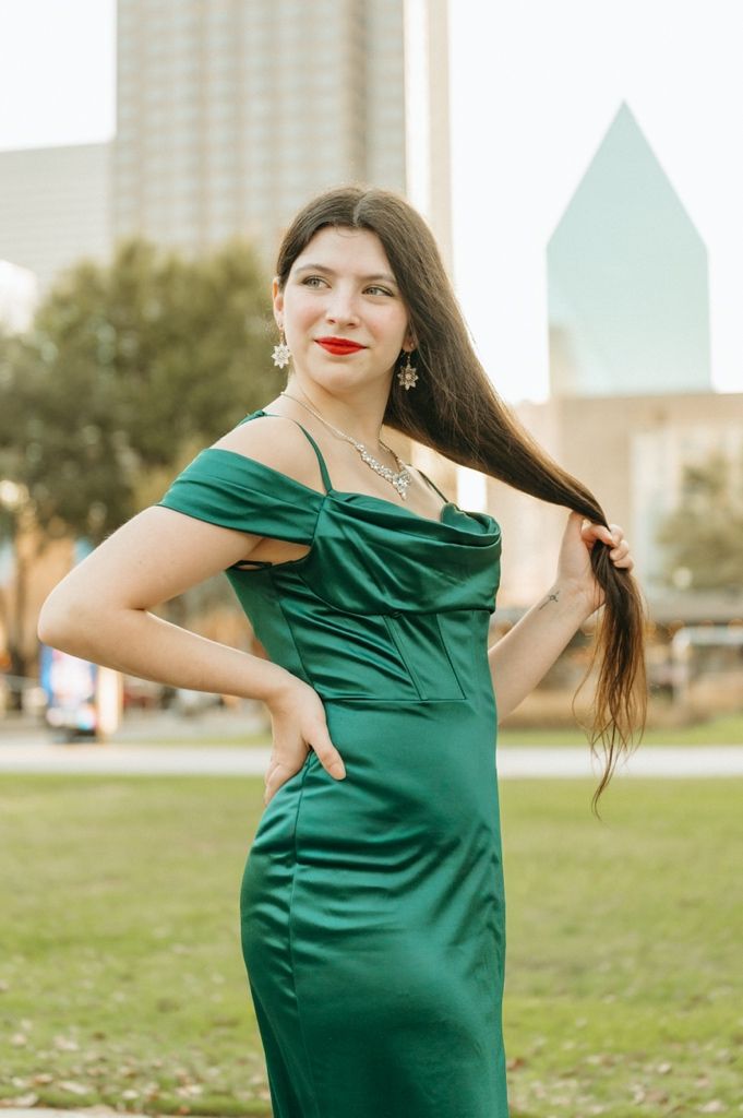 Woman in elegant green satin off-shoulder gown posing outdoors with Dallas skyline backdrop.