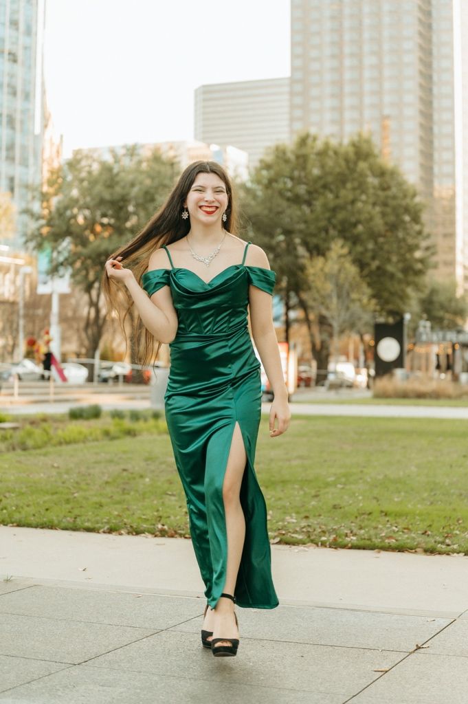 Woman in elegant emerald green satin off-shoulder gown with slit walks confidently in urban park setting.