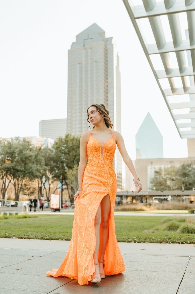 Woman in orange sequin slit gown poses outdoors with Dallas skyline skyscrapers in background.