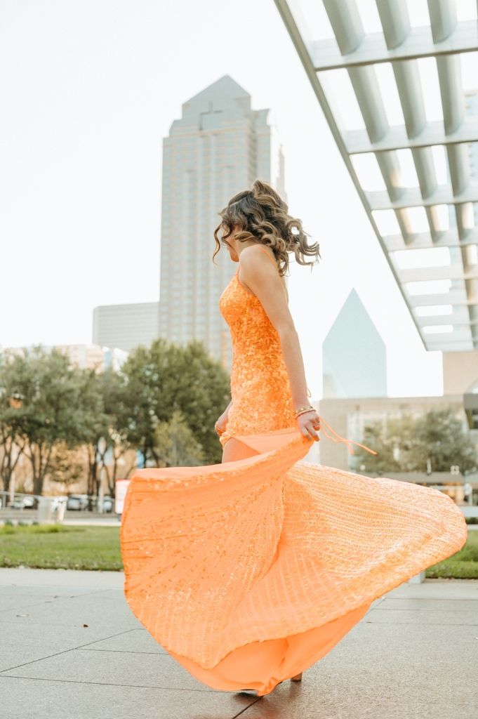 Young woman twirling in an orange sequin gown outdoors with city skyline in the background.