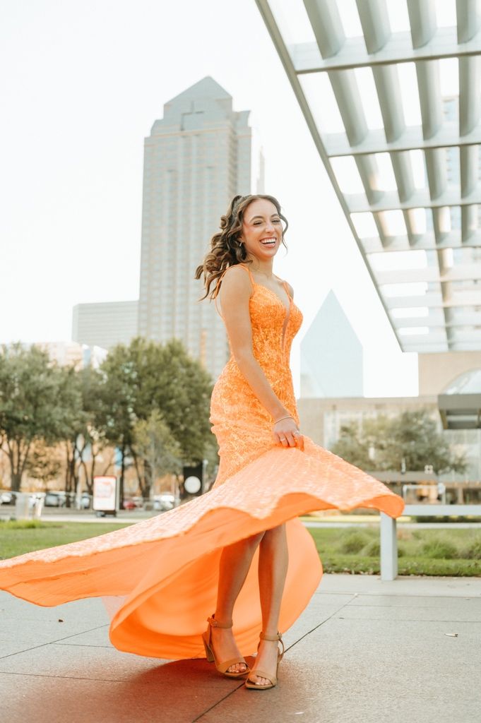 Young woman twirling in an orange lace mermaid gown outdoors, with a city skyline in the background.