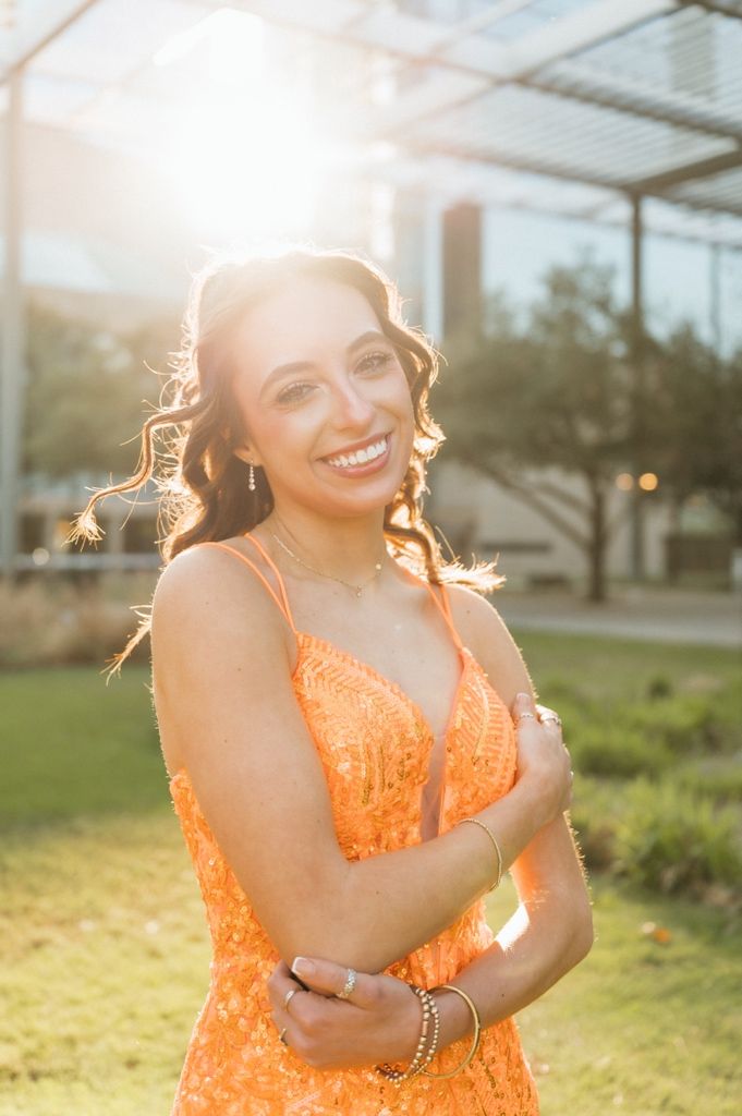 Young woman in orange sequin dress smiling outdoors with golden sunlight backdrop.