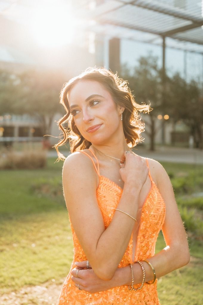 Young woman in orange sequin gown poses outdoors at golden hour with sun flare behind her.