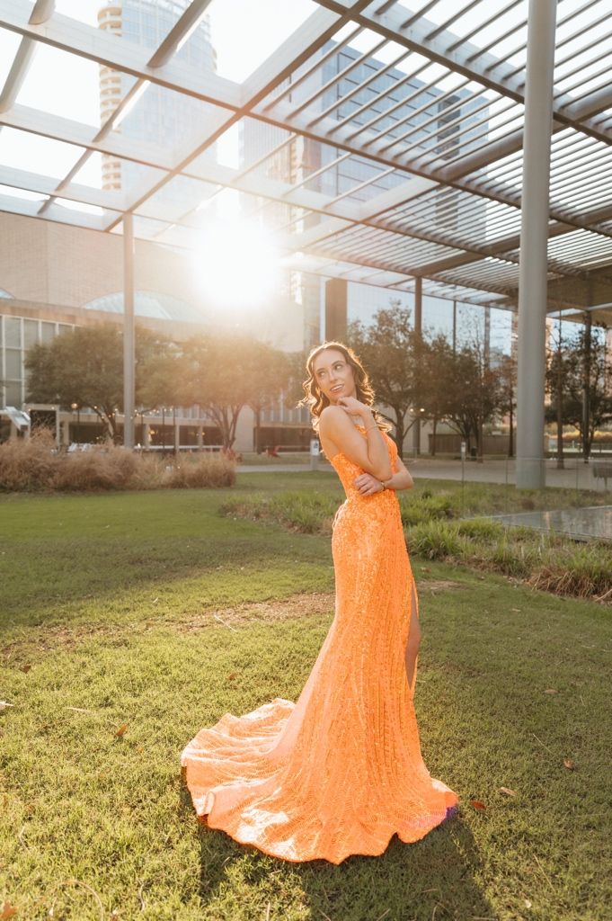 Elegant woman in orange sequin gown with train poses in sunlit outdoor venue at golden hour.