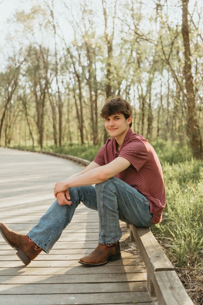 Young man in red striped polo and jeans sitting casually on a curved wooden boardwalk in nature.