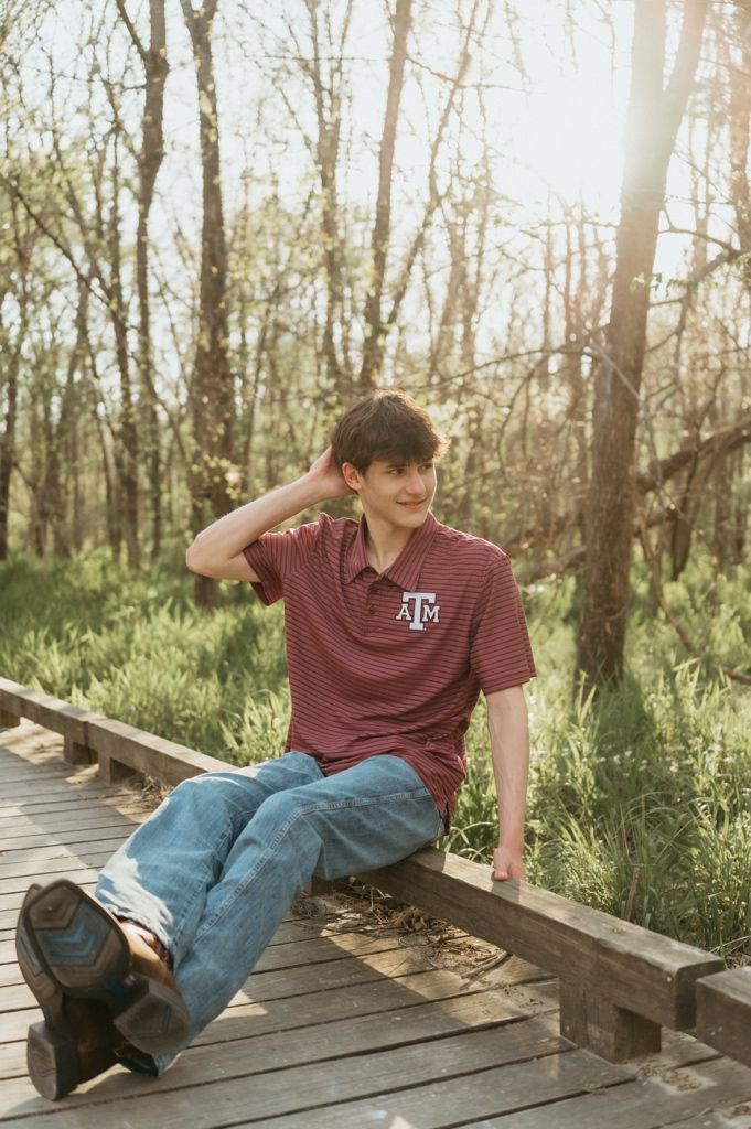 Teen boy in Texas A&M polo sits on wooden bridge railing surrounded by lush green trees in golden hour light.