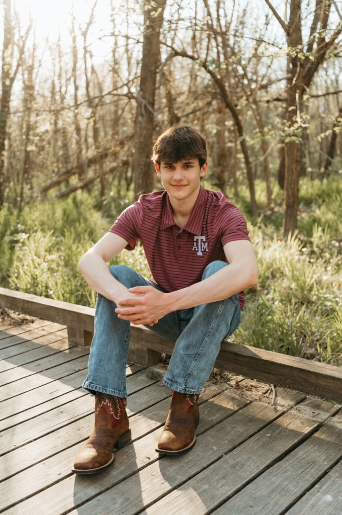 Young man in Texas A&M polo shirt and cowboy boots sitting on wooden deck outdoors.