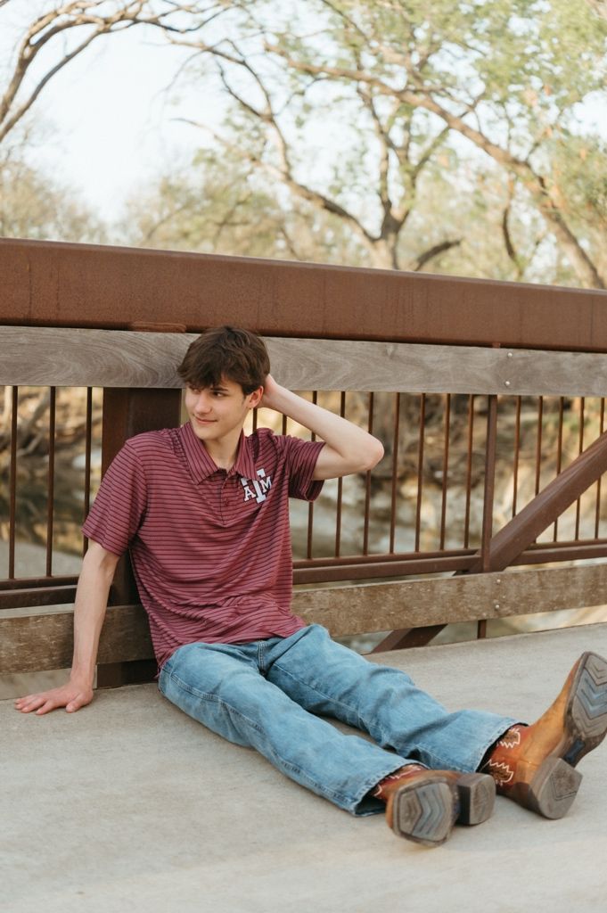 Teen boy in Texas A&M polo sits on bridge deck in jeans and boots, gazing thoughtfully to the side.