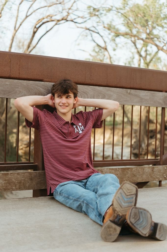 Teen boy in Texas A&M polo sits relaxed on bridge deck with hands behind head, wearing jeans and cowboy boots.