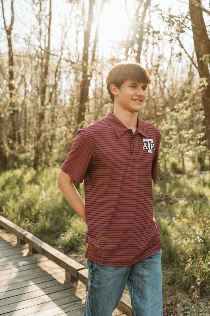 Young man in Texas A&M maroon striped polo and jeans smiling on a wooden boardwalk surrounded by trees.