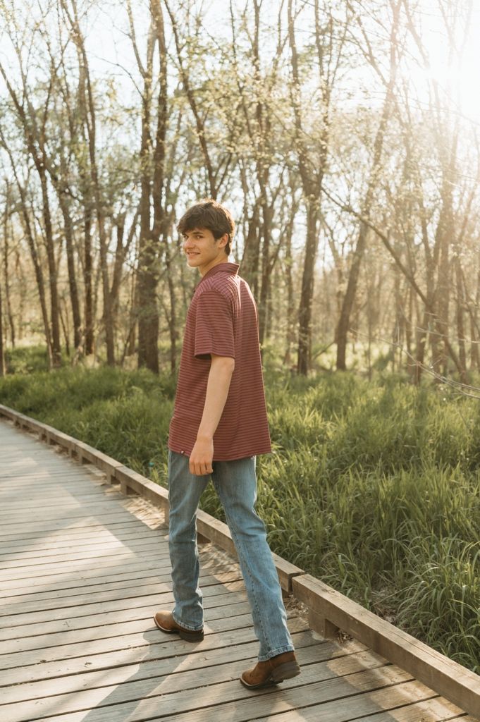 Young man in red shirt and jeans walking on wooden boardwalk path through lush green forest in sunlight.