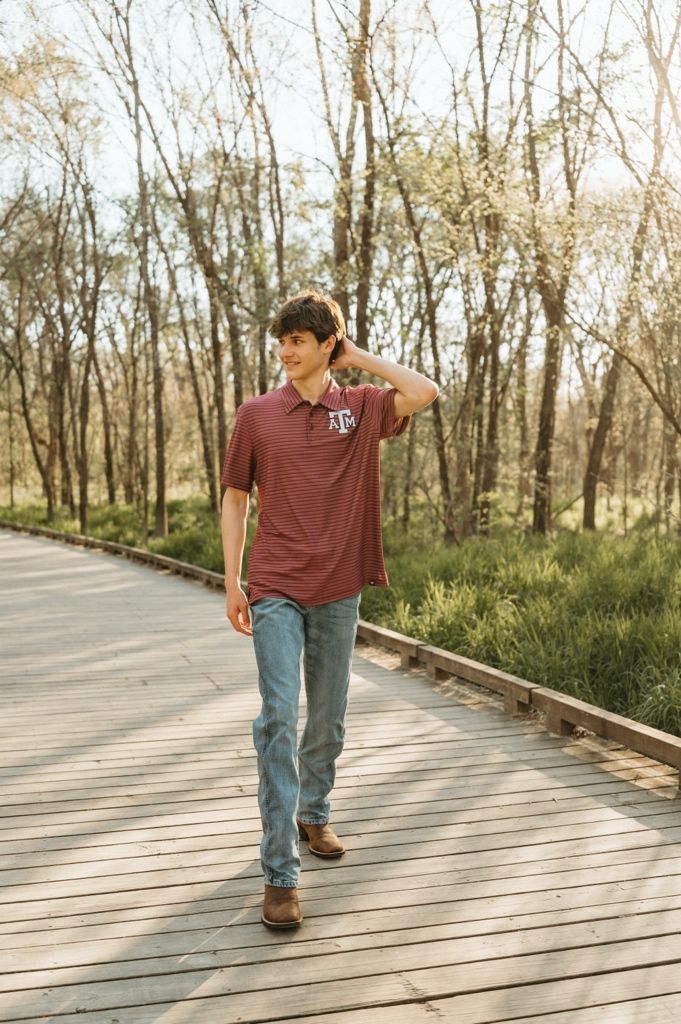 Teen boy in Texas A&M maroon polo and jeans walking on a wooden boardwalk path through a wooded area.