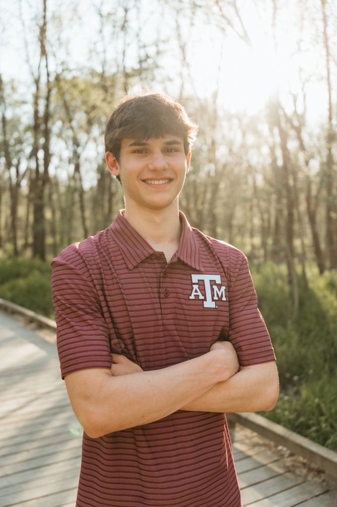 Young man in Texas A&M maroon polo shirt smiling with arms crossed on a sunny wooded boardwalk.