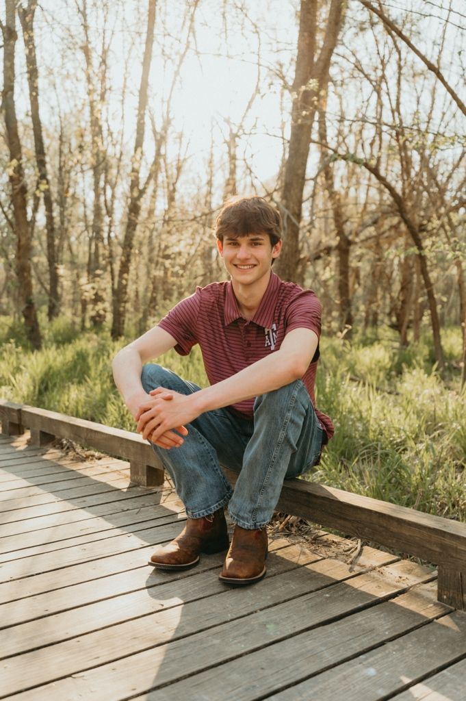 Young man in striped polo and jeans smiling on a wooden boardwalk surrounded by trees and greenery.