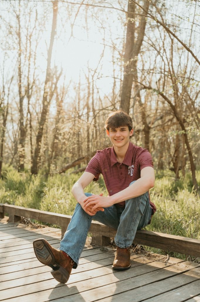 Smiling teen in Texas A&M polo and jeans sitting relaxed on a wooden boardwalk surrounded by trees.