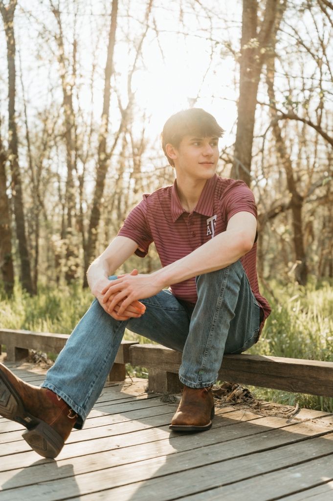 Young man in striped polo and jeans sitting on wooden deck surrounded by sunlit trees.