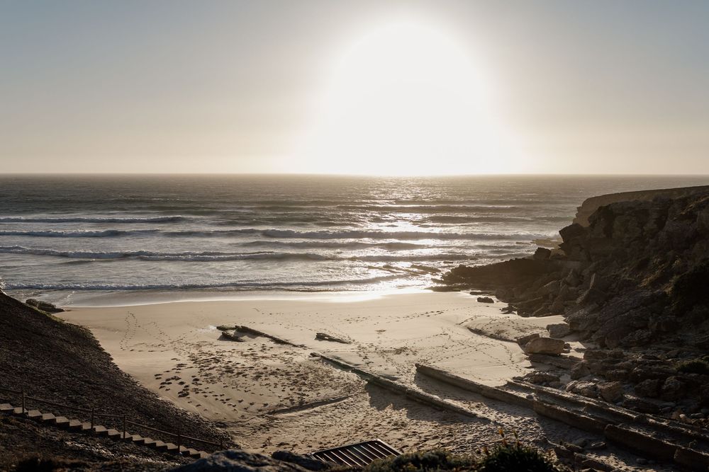 Sunset illuminates a dramatic coastal landscape with rocky cliffs and waves rolling onto a sandy beach.