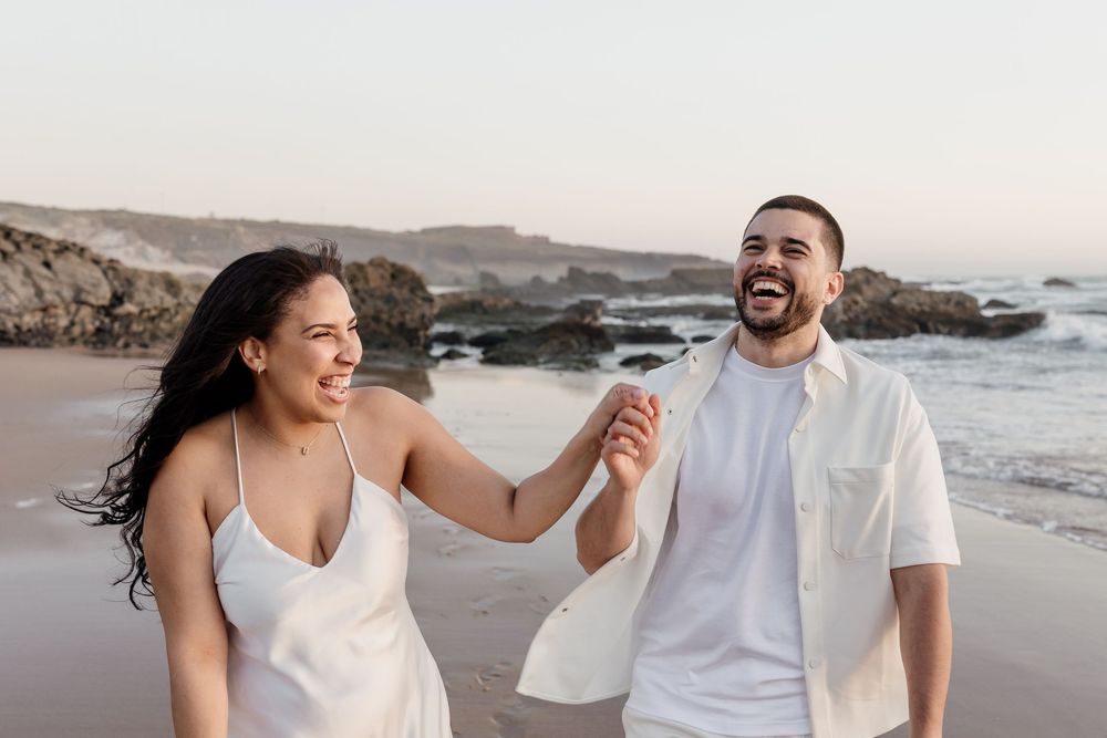 Happy couple holding hands and laughing while walking along a rocky beach at sunset.