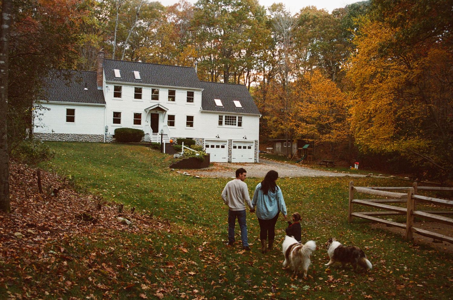 Family enjoying an autumn day outdoors near a white farmhouse surrounded by colorful fall foliage.