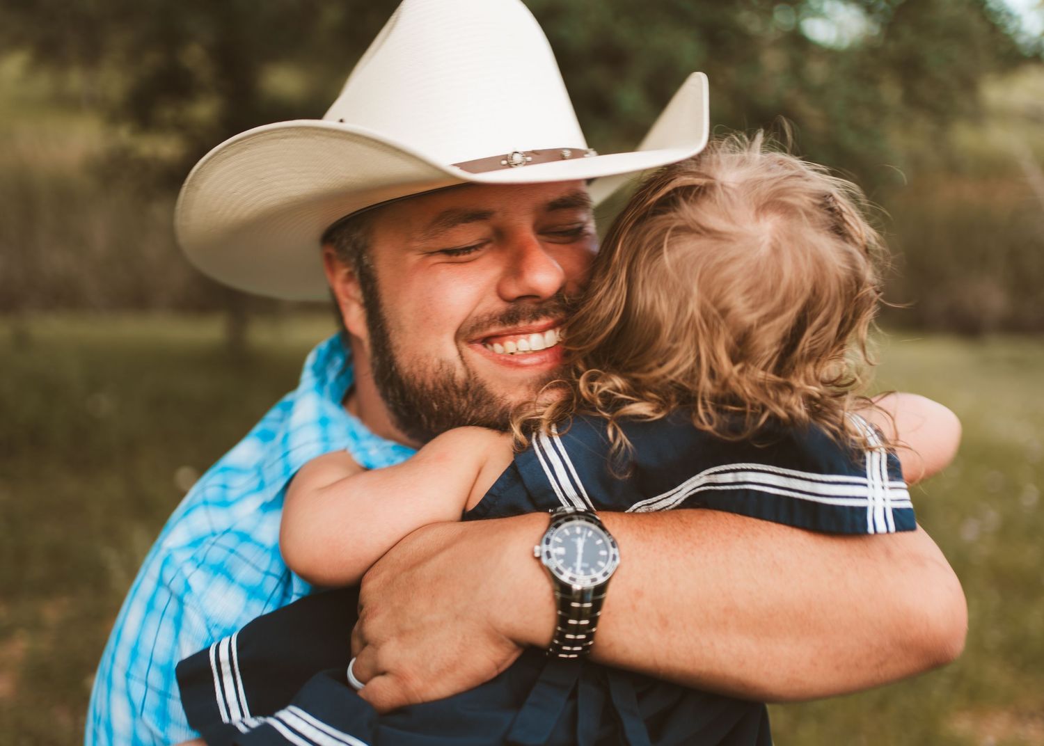 Smiling man in cowboy hat sharing a warm hug with a young child outdoors.