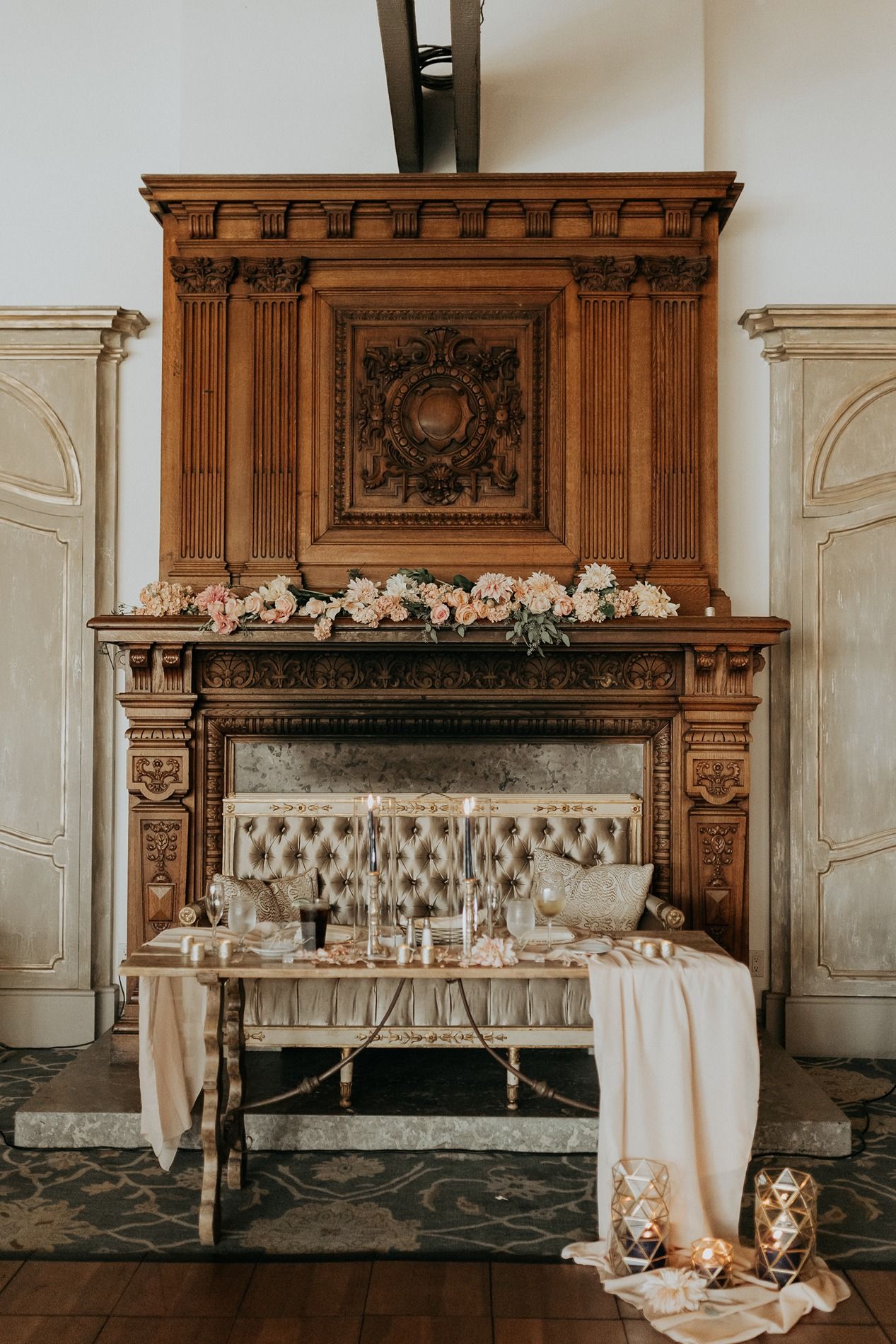 Elegant wedding sweetheart table set before ornate wooden fireplace mantel adorned with blush floral arrangements.