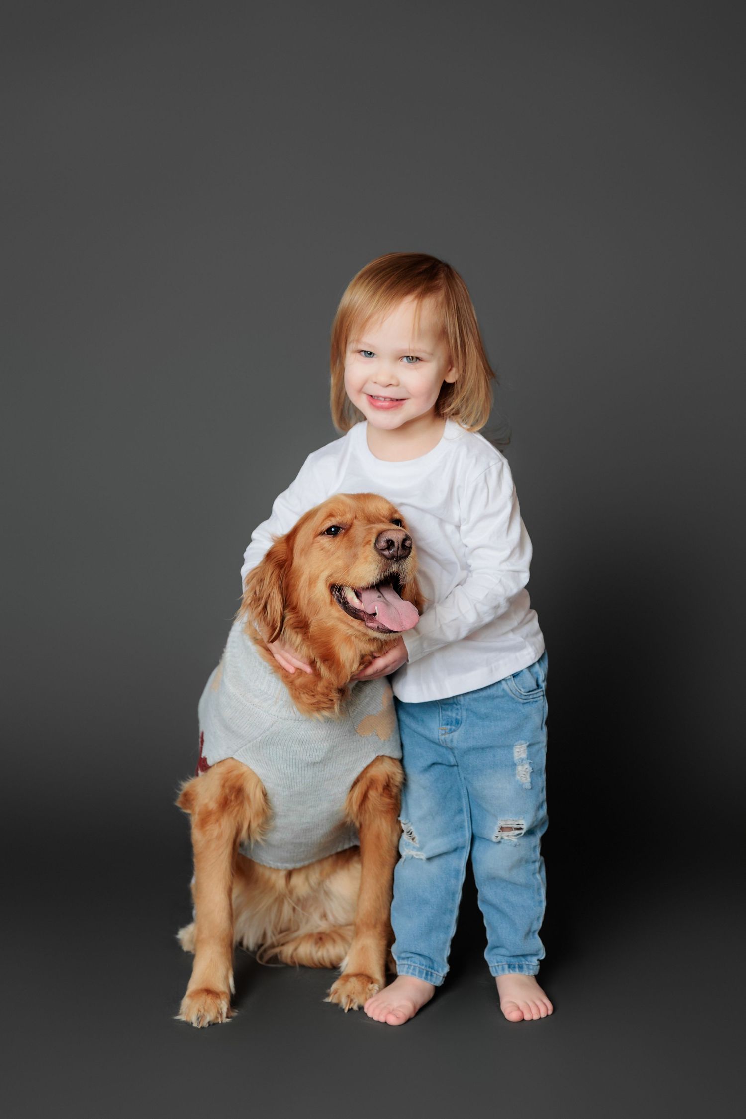 A golden retriever puppy sits beside a child in jeans and white shirt against a dark studio background.