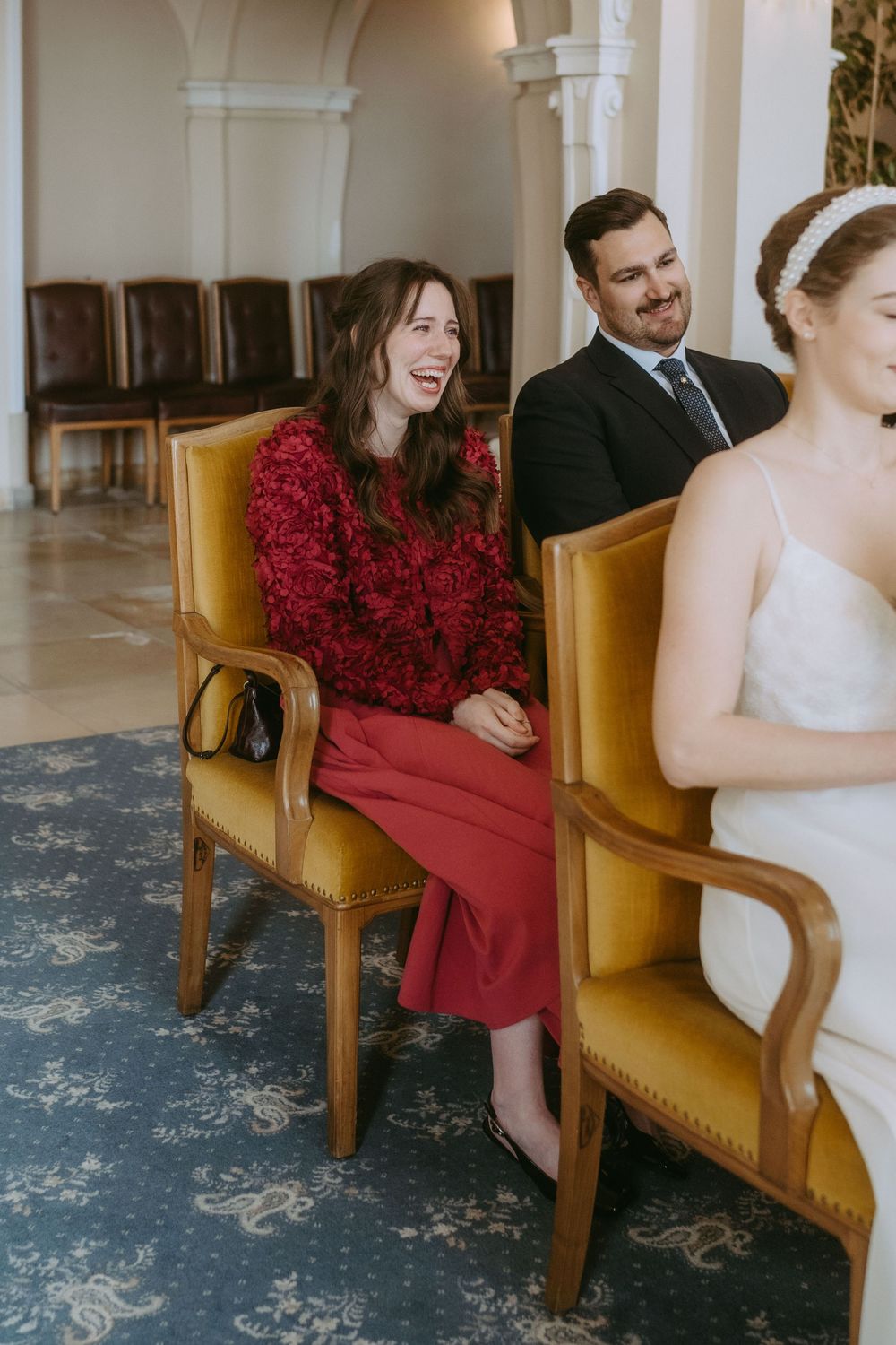 A wedding couple sits in elegant gold chairs with red floral arrangements in an ornate interior setting.
