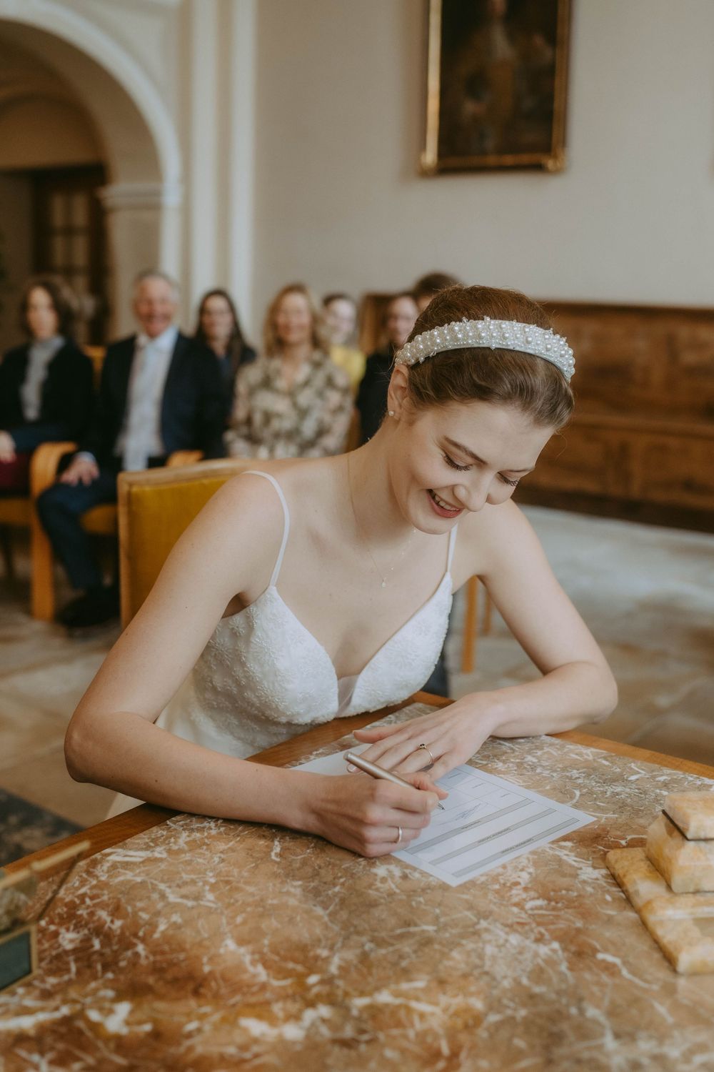A bride in a white dress signs documents at a marble table during a wedding ceremony.