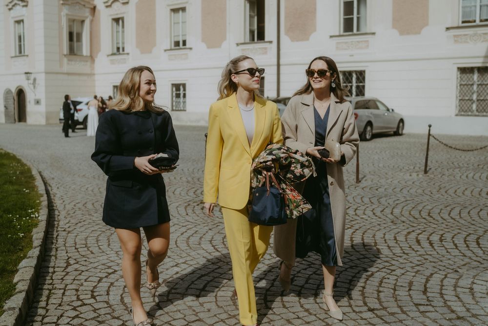 Three stylishly dressed friends walk together on a cobblestone street, with one wearing a striking yellow pantsuit.