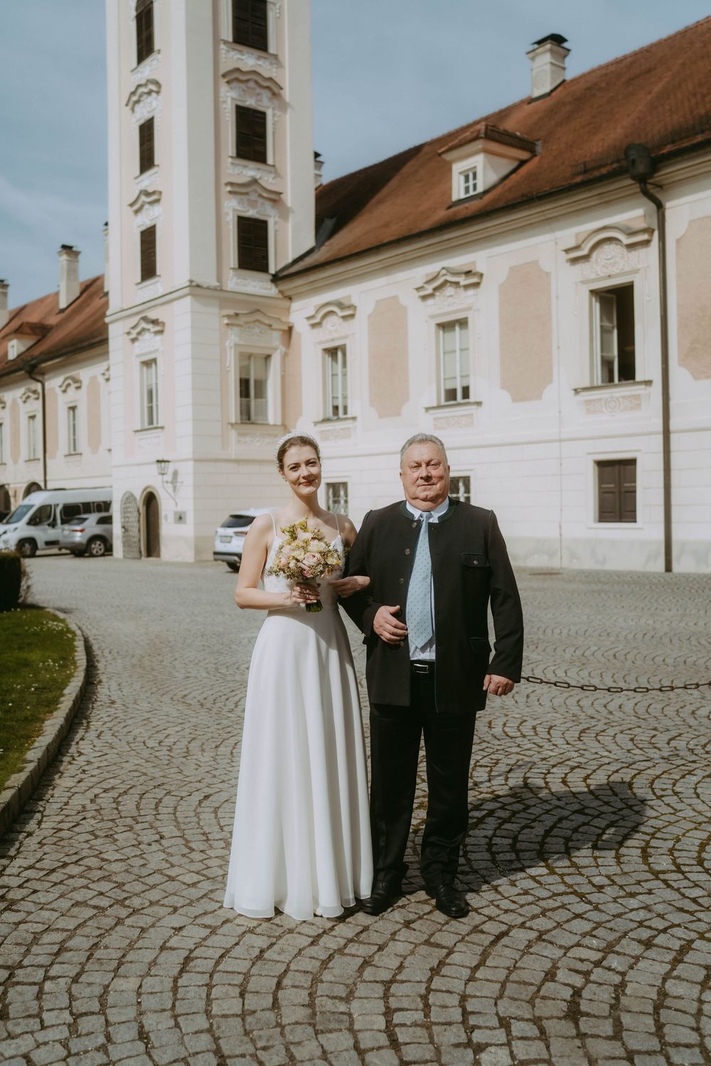 A wedding couple poses in front of a historic white church building with a tall bell tower and cobblestone courtyard.