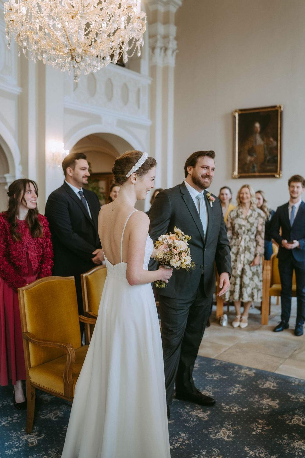 Elegant wedding ceremony in a historic venue with crystal chandeliers and vintage yellow chairs.